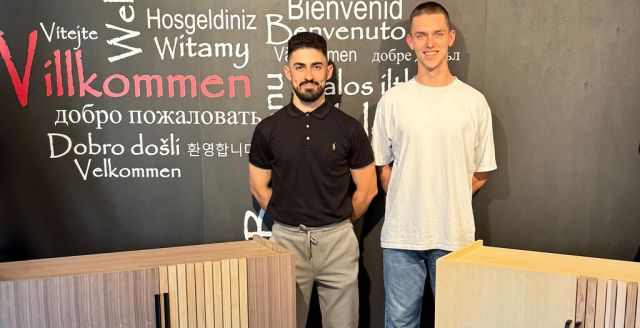 Two young men pose confidently beside modern cabinets against a multilingual welcome wall, showcasing craftsmanship.