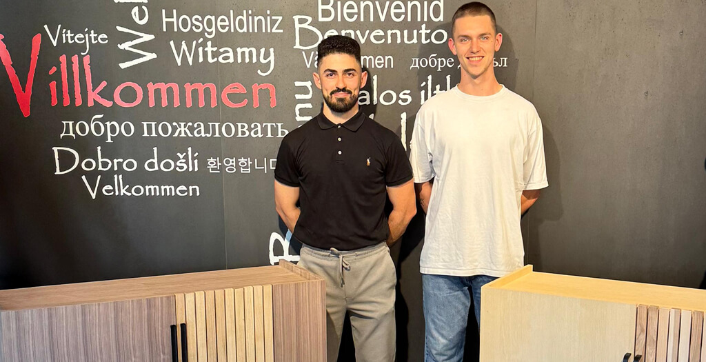 Two young men stand together in front of a welcoming multilingual mural, showcasing their partnership in craftsmanship.